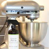 Peppermint Vanilla All Purpose Cleaner bottle on granite counter, in front of a silver stand-up mixer, safe and natural kitchen cleaner.