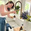 woman adding water into a  Red's Gone Green glass spray bottle that has lavender vanilla all purpose cleaner concentrate in it